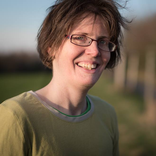 Porträtfoto von Melanie. Man sieht Melanie lächelnd und mit leicht windzerzausten Haaren mit einem hellgrünen Shirt bekleidet vor einem unscharfen Hintergrund aus Wiese, Wald und etwas blauem Himmel.